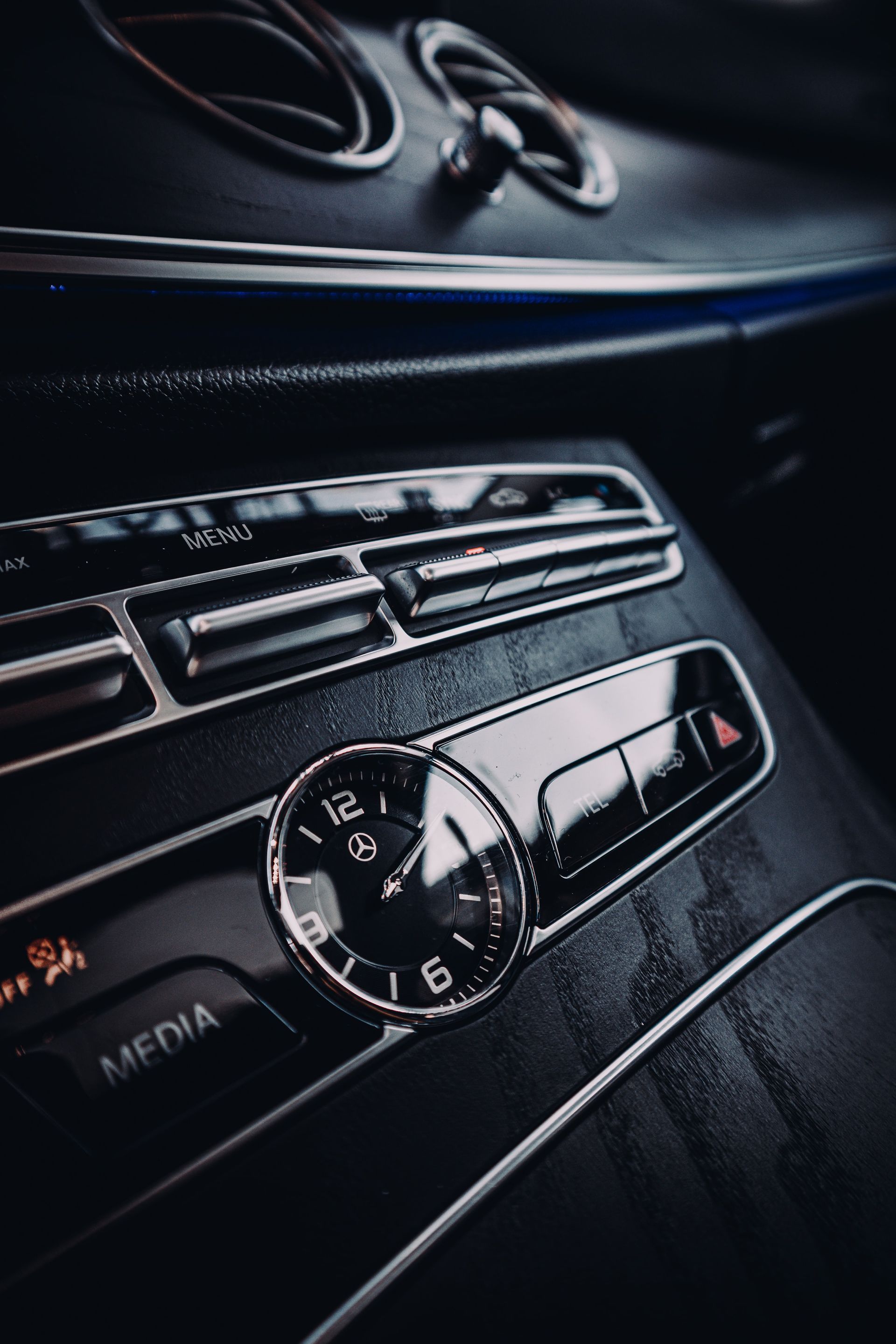 Close-up of a Mercedes-Benz car interior, showing a clock, air vents, and media control panel in dark tones.