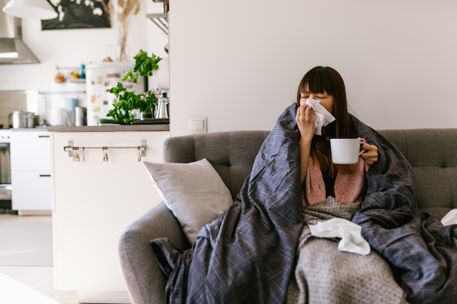 A woman is sitting on a couch with a cup of coffee and blowing her nose.