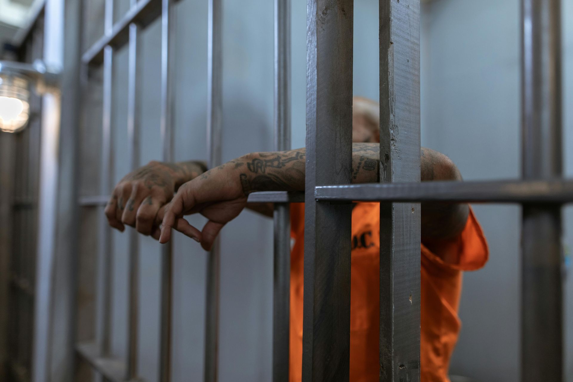 Person in orange prison uniform, hands on bars, behind bars.