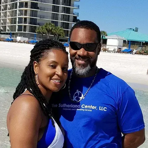 Smiling couple posing on a sunny beach, with a hotel in the background.