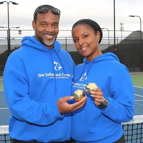 Two people in blue hoodies smiling and holding gold medals on a tennis court