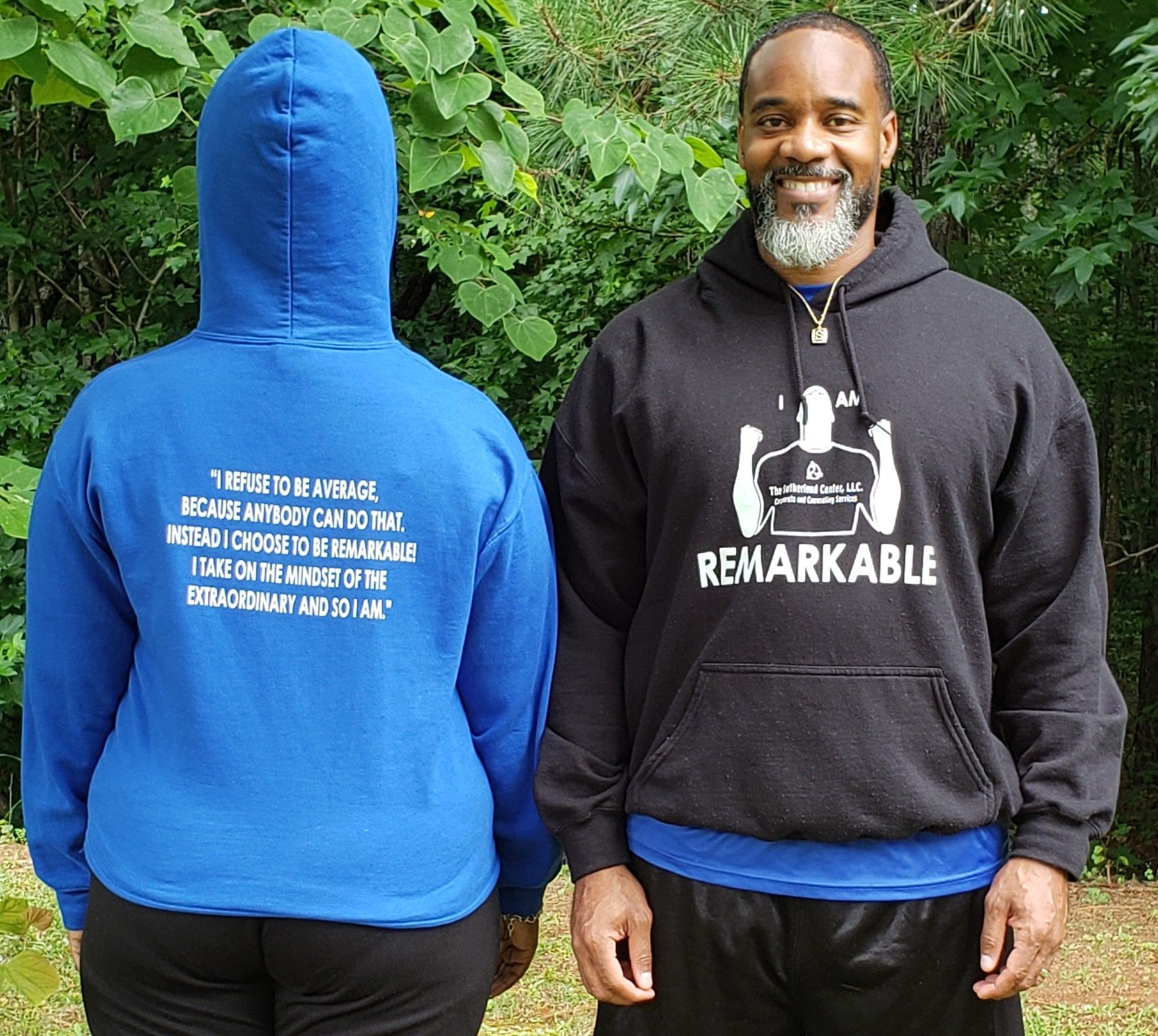 Two people wearing hoodies stand outdoors in front of green foliage; one faces away, the other smiles.