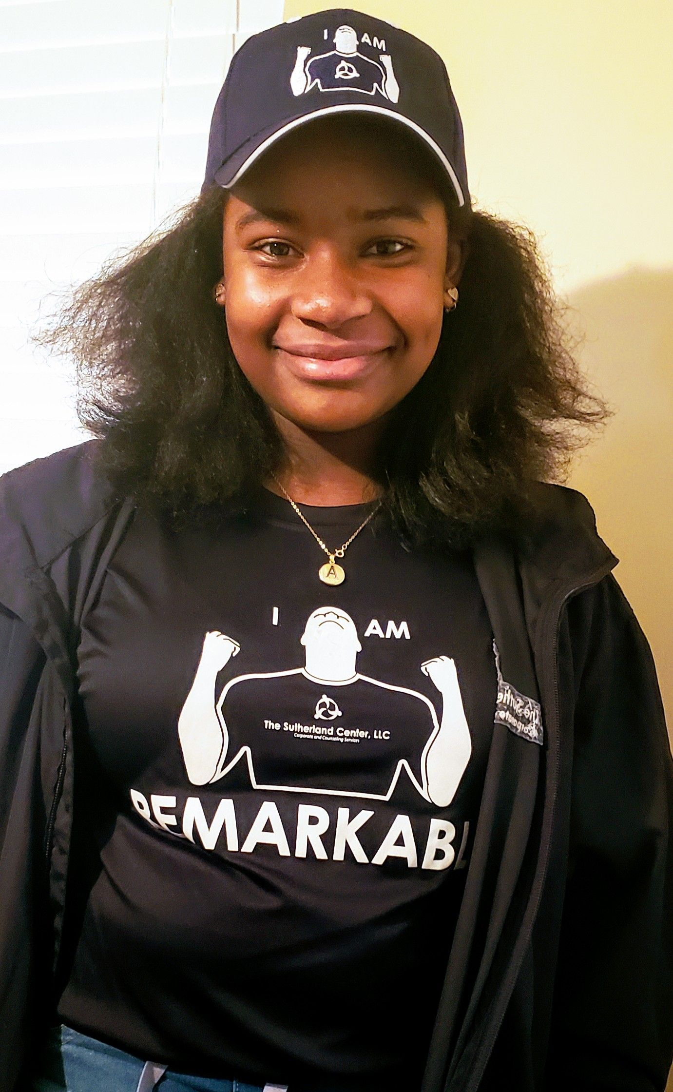 Smiling person in black cap and jacket, wearing a black graphic T-shirt against a light background.