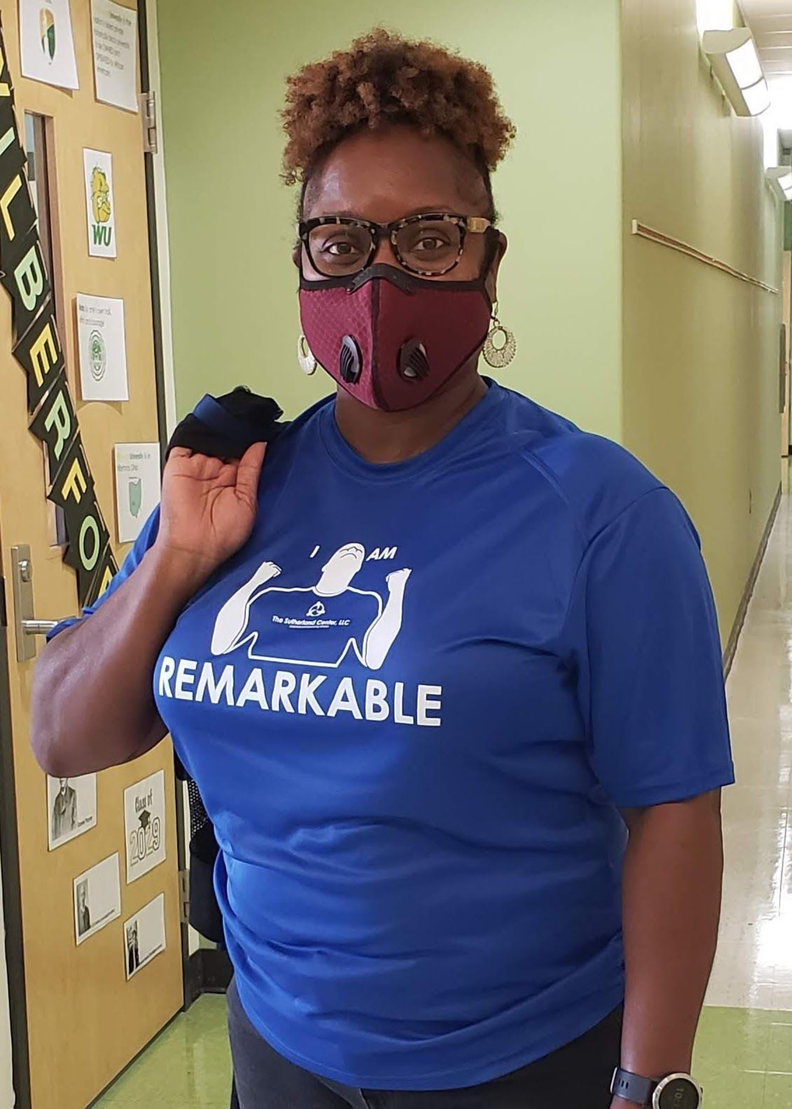 Woman in blue “REMARKABLE” shirt and maroon mask, standing in a hallway and holding a bag over her shoulder