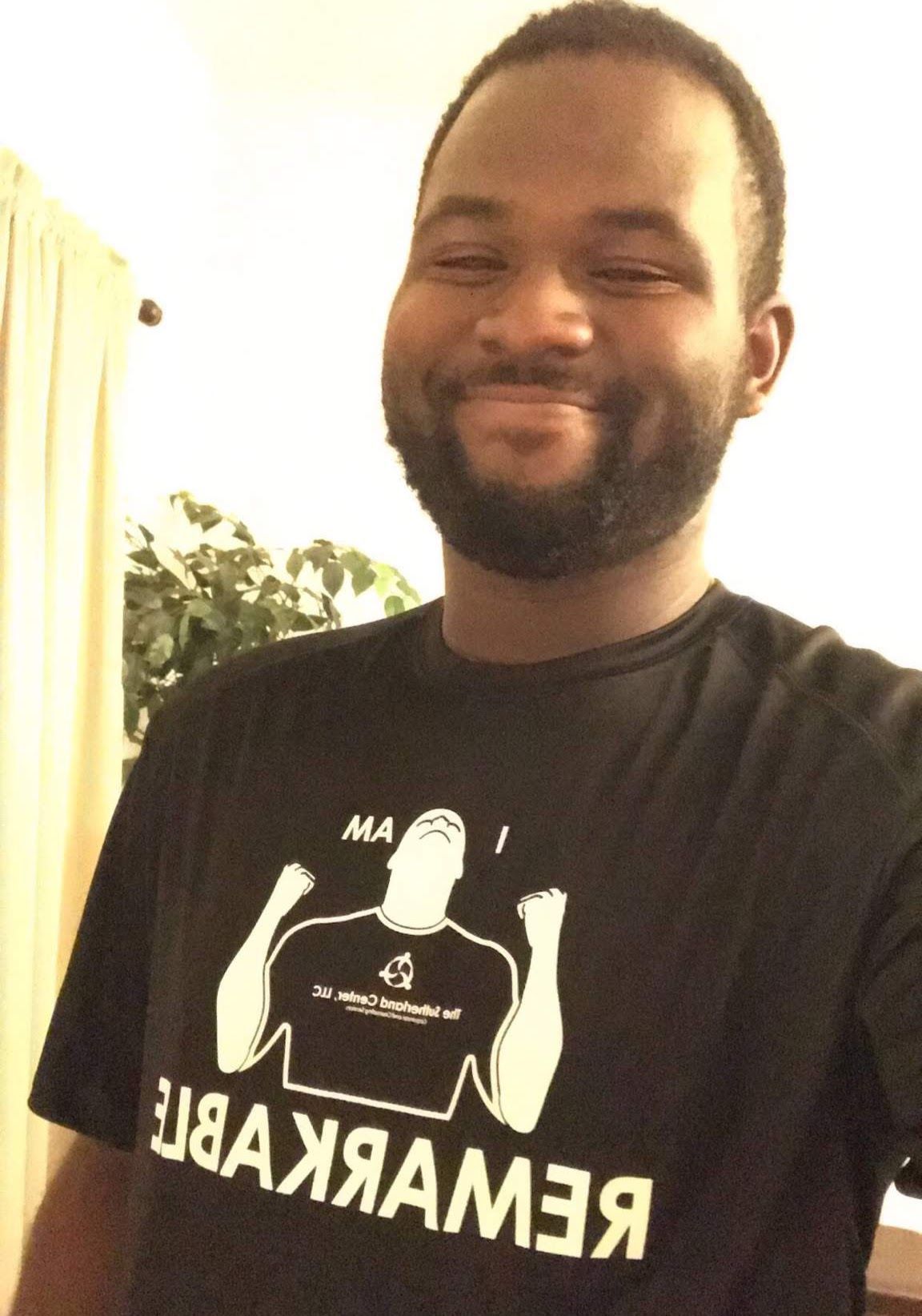 Smiling man in a black T-shirt standing indoors, with bright light and a plant in the background