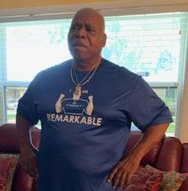 Man standing indoors in a navy “REMARKABLE” T-shirt, hands on hips, in front of a bright window.