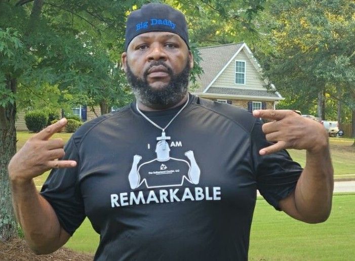 Man standing outdoors on a lawn, wearing a black “REMARKABLE” shirt and blue cap, making hand gestures.