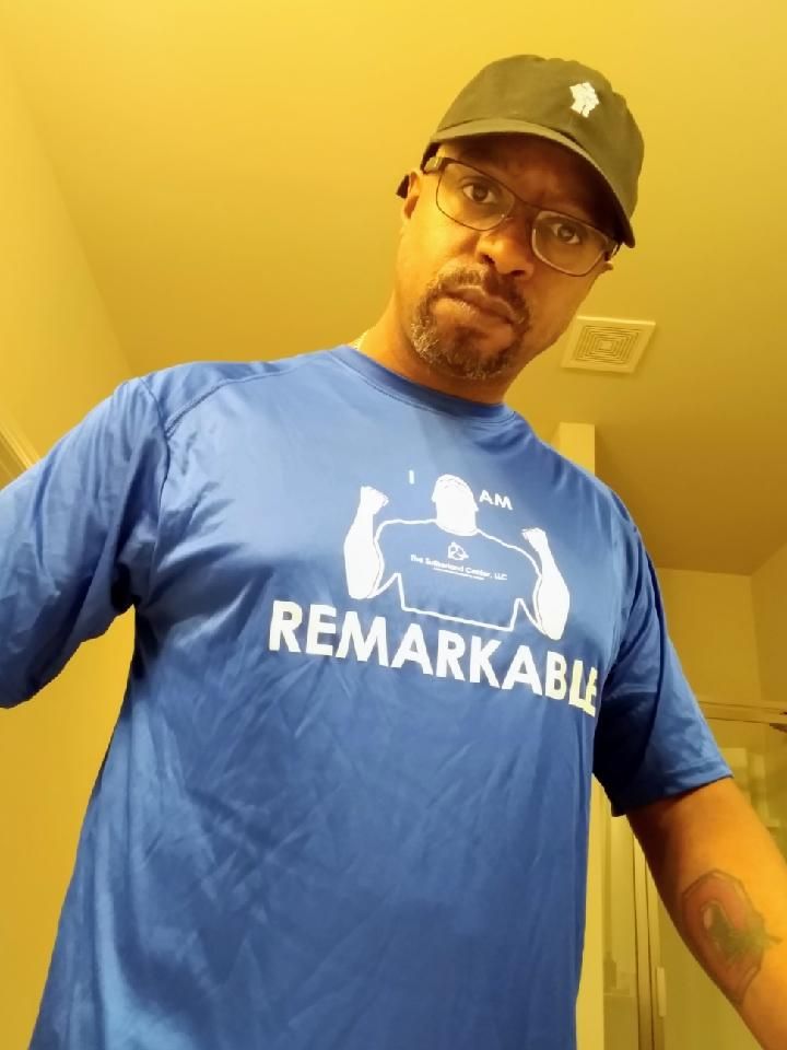 Person wearing a blue “REMARKABLE” shirt and black cap, standing indoors against a yellow wall.