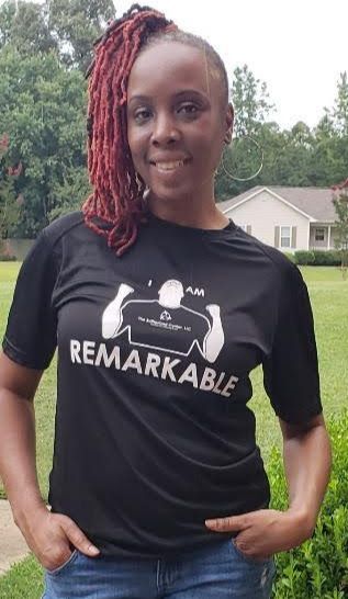 Woman outdoors in a black “Remarkable” T-shirt, standing on grass and smiling.