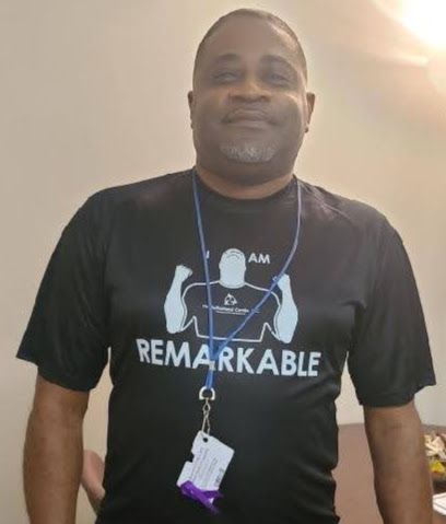 Man in a black “REMARKABLE” T-shirt and lanyard, standing indoors against a light wall.