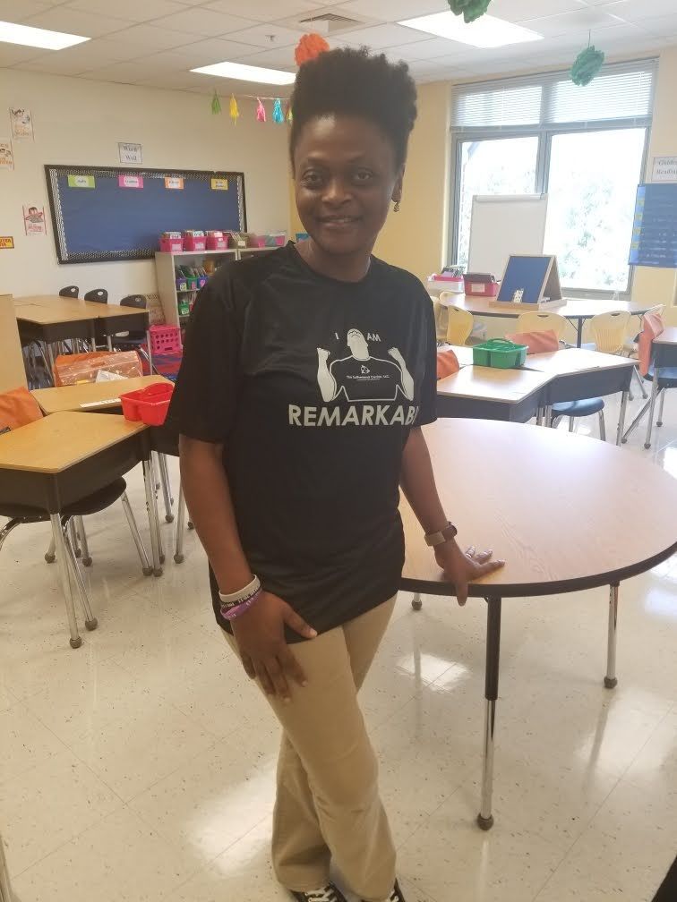 Person standing in a classroom beside a round table, wearing a black T-shirt and tan pants.