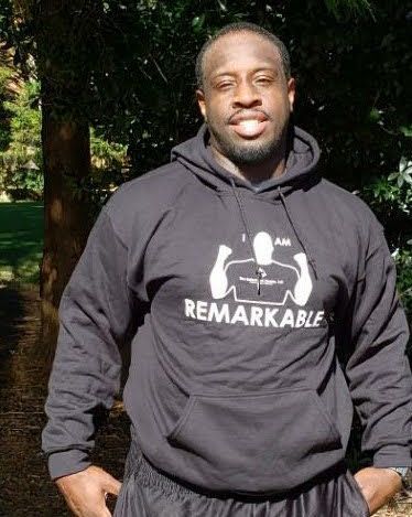 Person in a black hoodie with “I AM REMARKABLE” text, standing outdoors in sunlight.