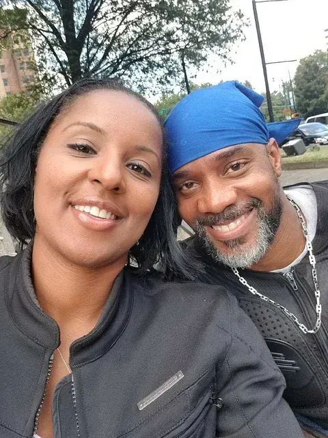 Two people smiling for a selfie outdoors, one in a black jacket and one in a blue headscarf.