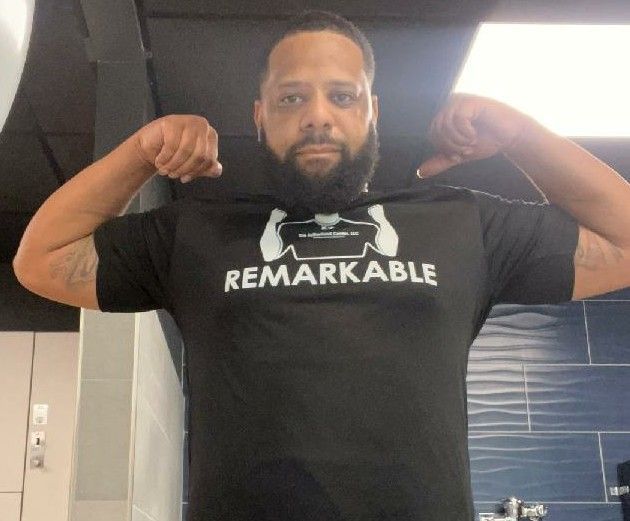 Man flexing both arms in a black “REMARKABLE” T-shirt indoors
