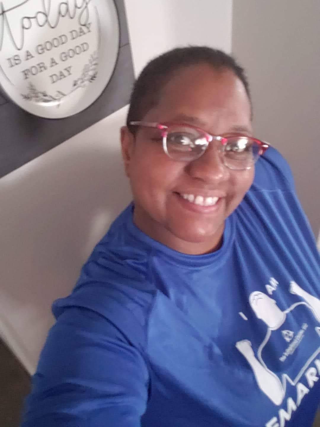 Smiling person in glasses and a blue shirt taking a selfie indoors.