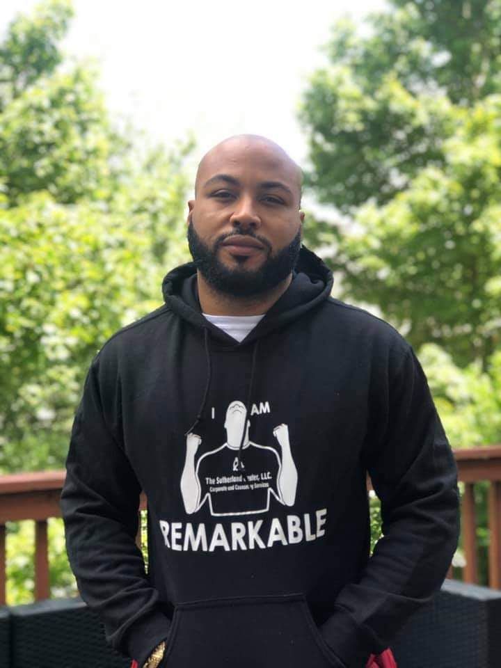 Person standing outdoors in a black hoodie with “I AM REMARKABLE” text, backed by green trees.