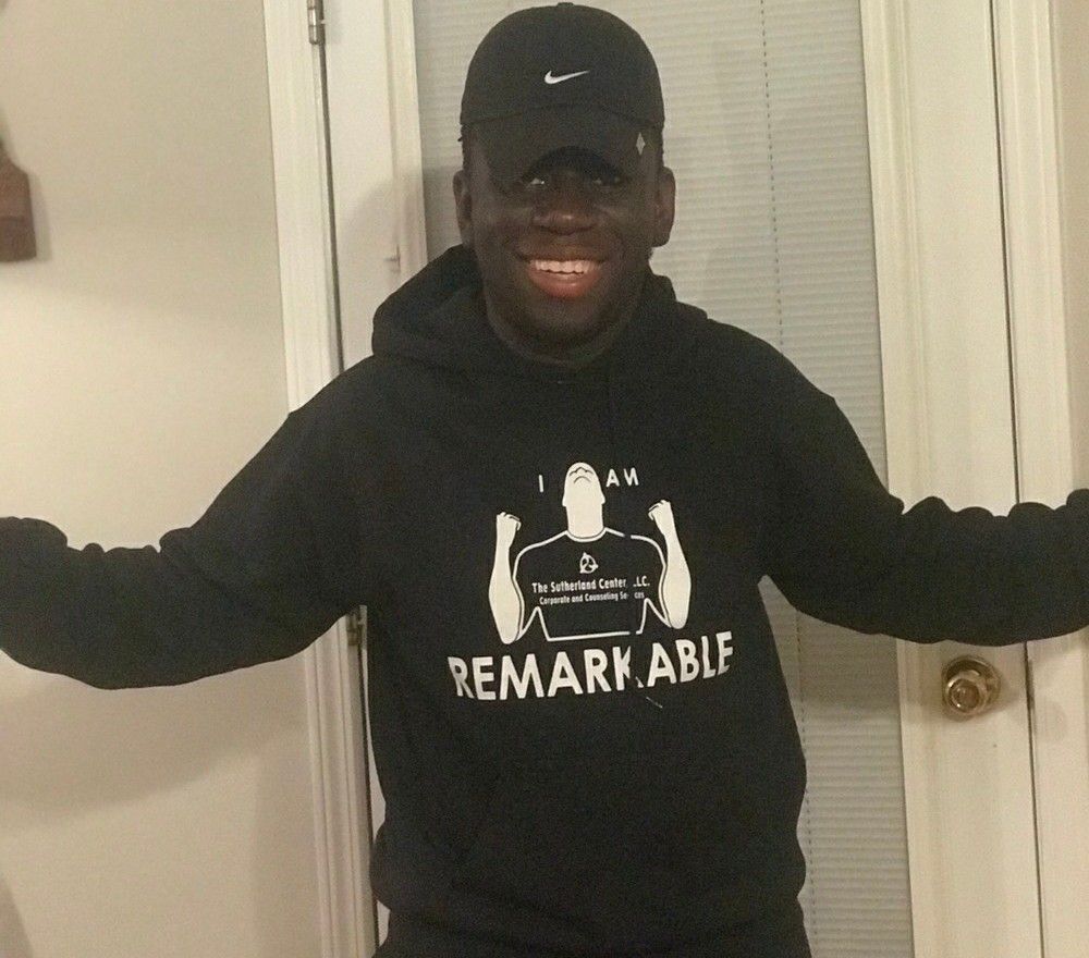 Person in black hoodie and cap stands smiling with arms out in a doorway.