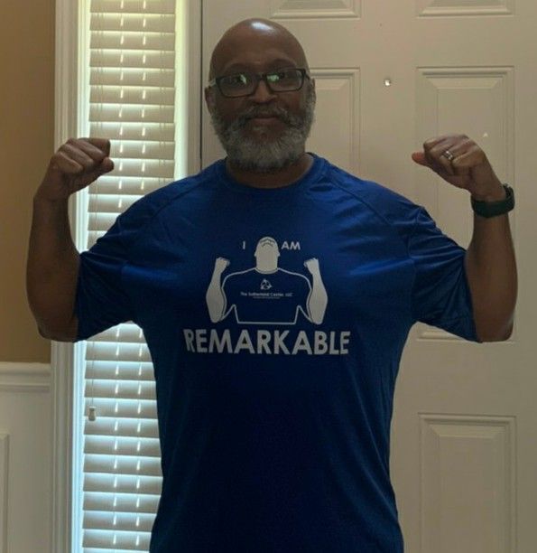Man in blue “REMARKABLE” shirt flexing both arms indoors, standing in front of a door.