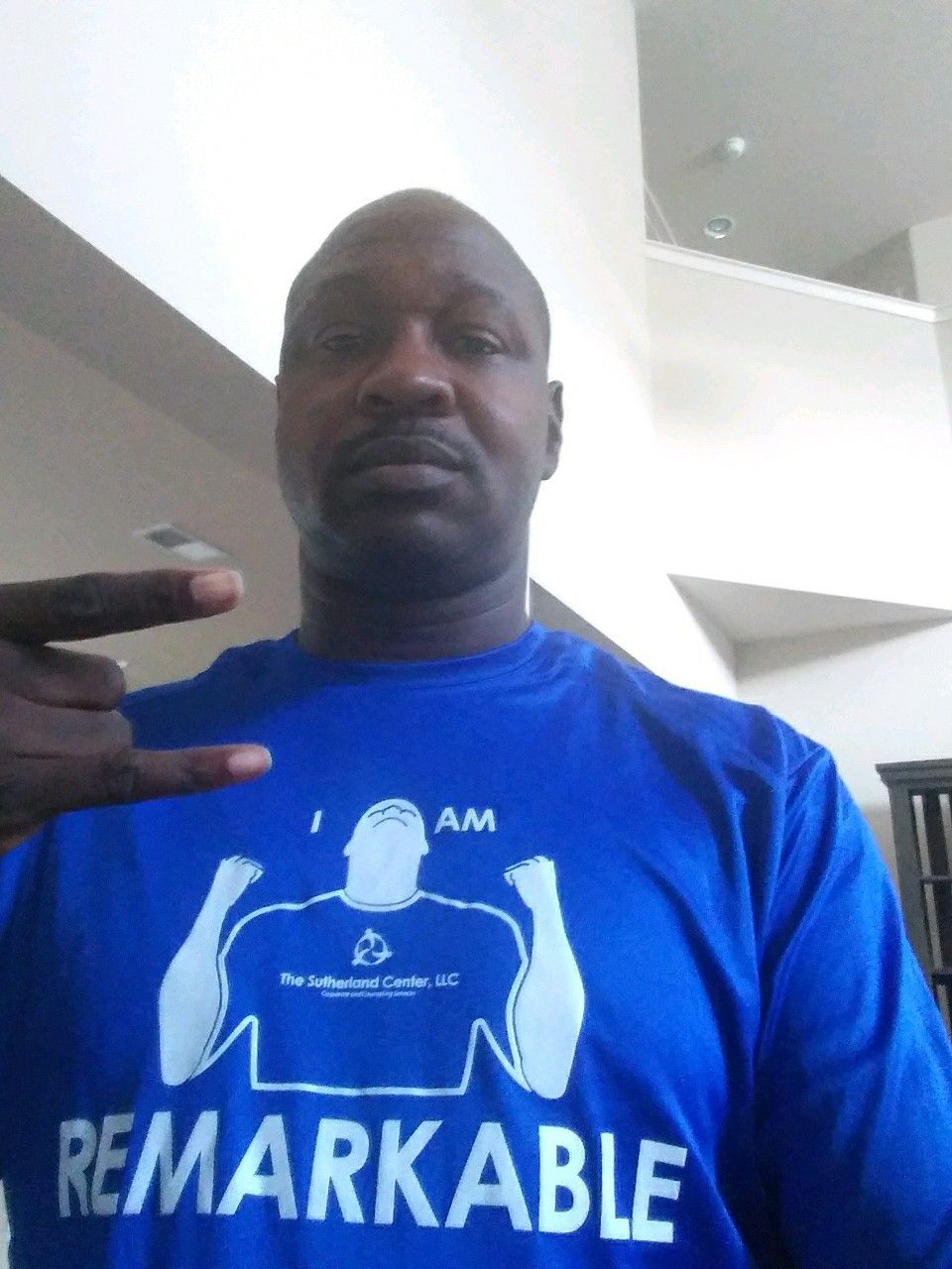 Man in blue REMARKABLE shirt making a hand gesture indoors, standing against a white staircase.