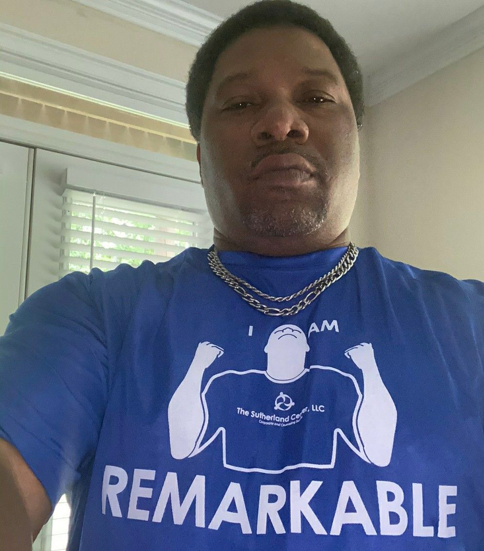 Person in a blue “I AM REMARKABLE” shirt and gold chains taking a selfie indoors.