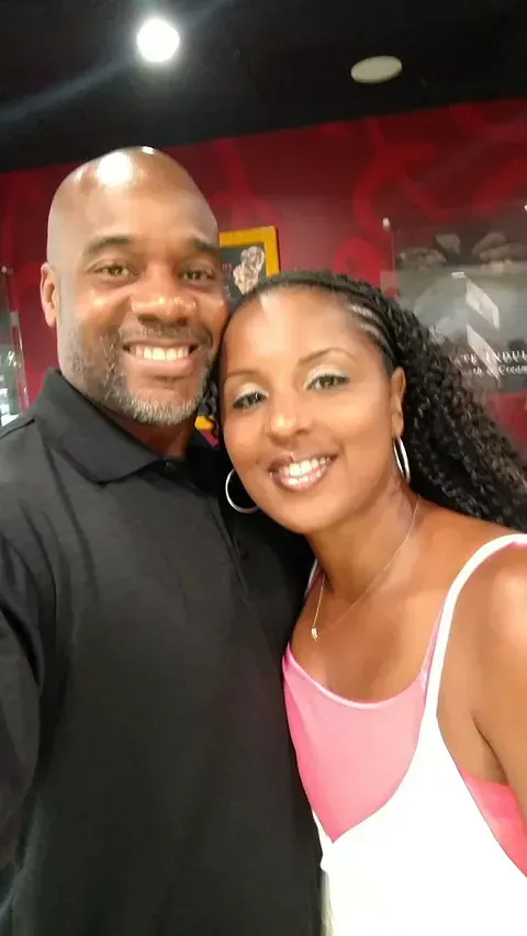 Two people smiling for a selfie indoors, with a red wall and ceiling lights in the background.