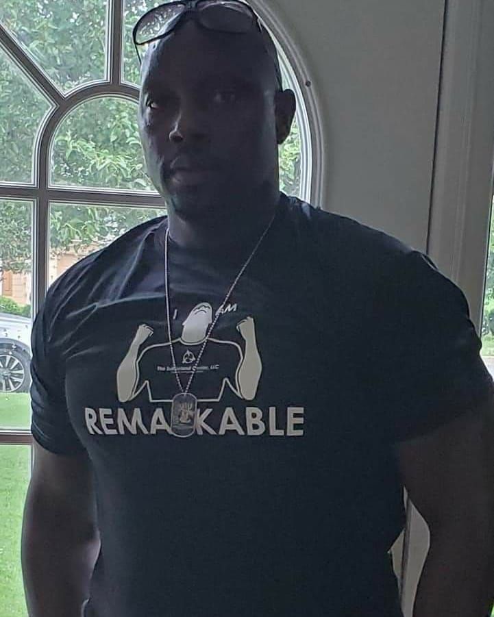 Man standing indoors by a window, wearing a black “REMARKABLE” T-shirt and earphones.