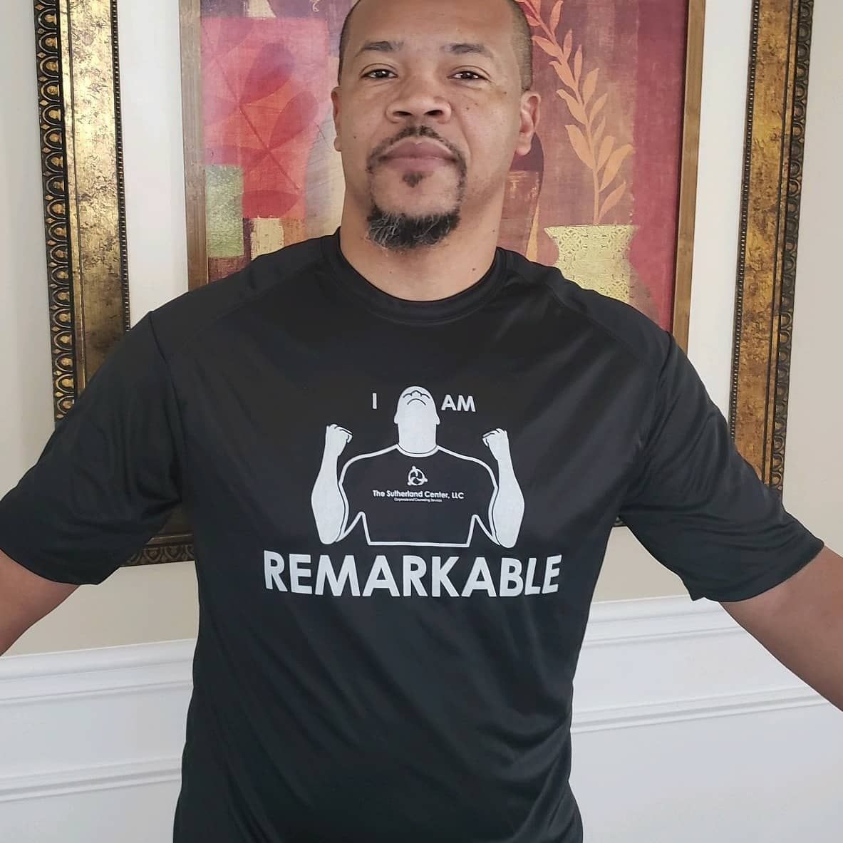 Man wearing a black “Remarkable” T-shirt, standing indoors with arms outstretched in front of framed wall art.