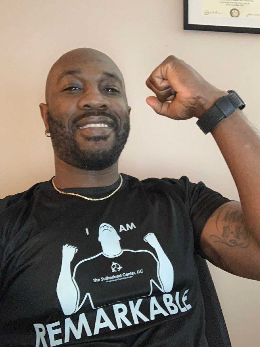Smiling man in black T-shirt flexing his arm indoors, with a framed certificate on the wall behind him.