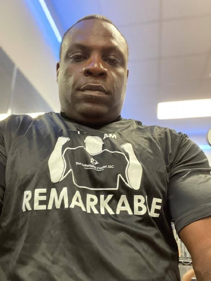 Man wearing a black “REMARKABLE” T-shirt, seated indoors under bright ceiling lights.