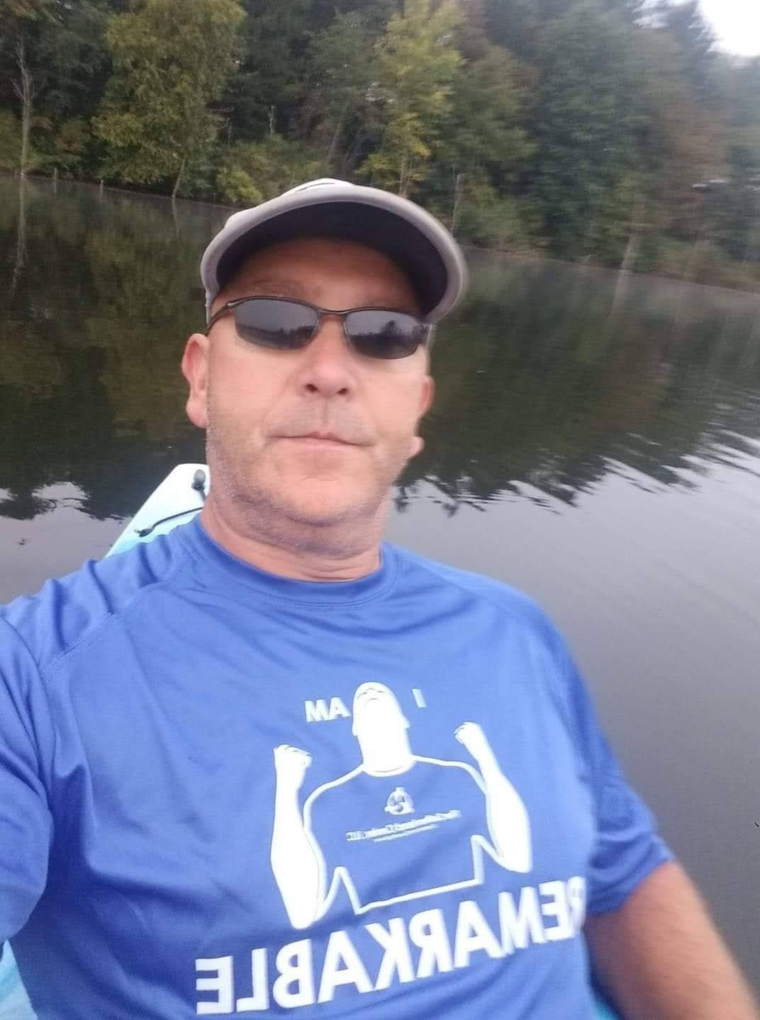 Man in sunglasses and cap sitting in a kayak on a calm lake with wooded shoreline.