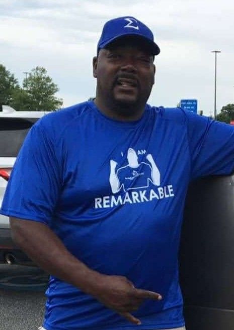 Man in blue “Remarkable” T-shirt and cap standing outdoors beside a vehicle, pointing downward.