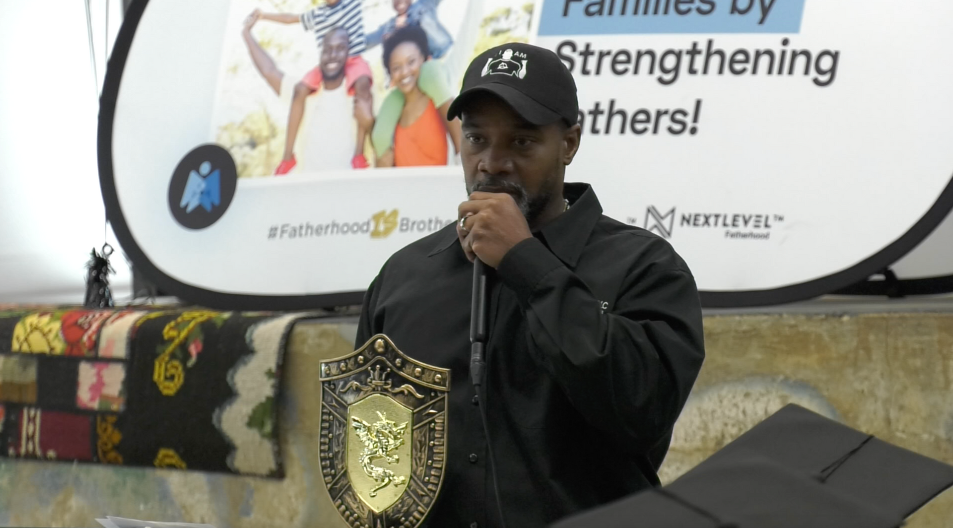 Person speaking at a podium with a gold championship-style belt, in front of a banner reading “Families by strengthening others!”
