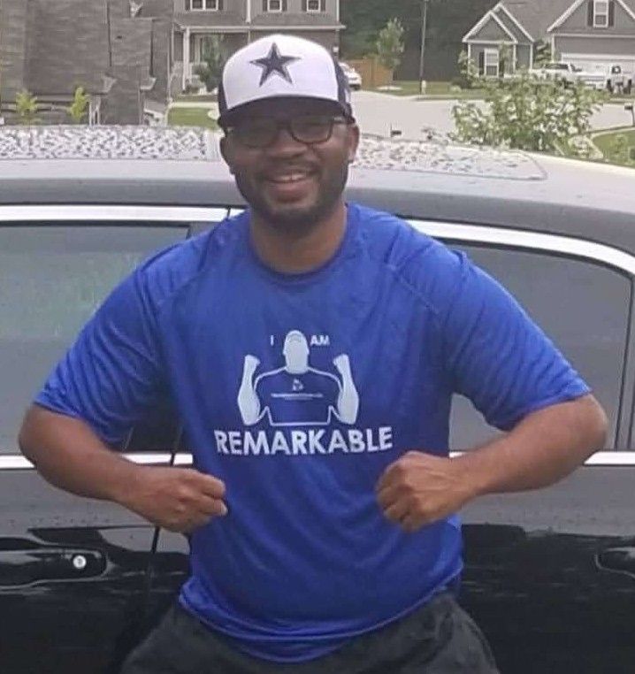 Man smiling and flexing in blue “REMARKABLE” shirt beside a car in a neighborhood driveway