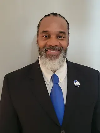 Smiling person in a black suit with a blue tie and lapel pin against a plain light wall