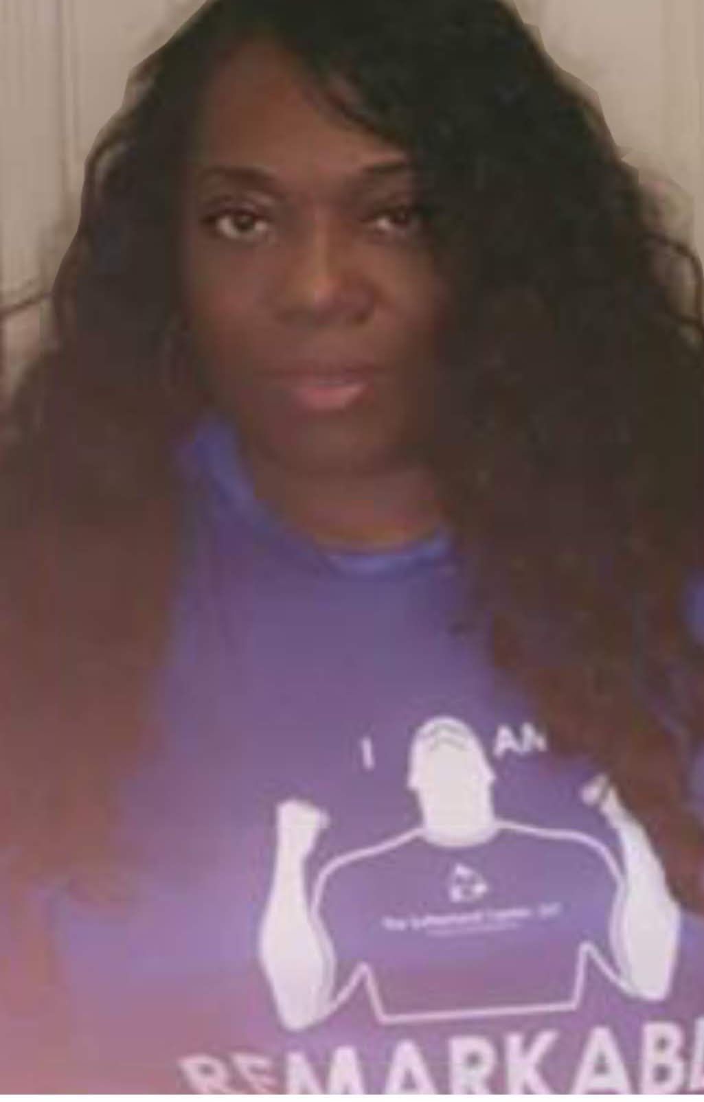 Person with long dark hair wearing a purple shirt, facing the camera indoors.