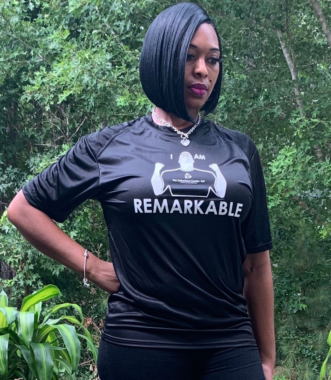Woman standing outdoors in a black “REMARKABLE” T-shirt, hands on hips, against green foliage.