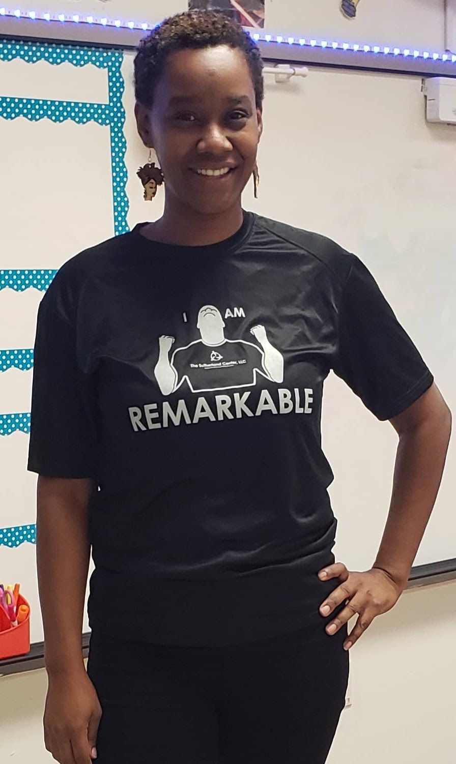 Person standing indoors, smiling, wearing a black “REMARKABLE” T-shirt with hands on hips.