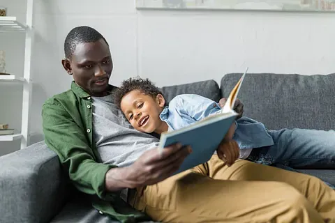 Two people relax on a gray couch, reading a blue book together in a bright living room.
