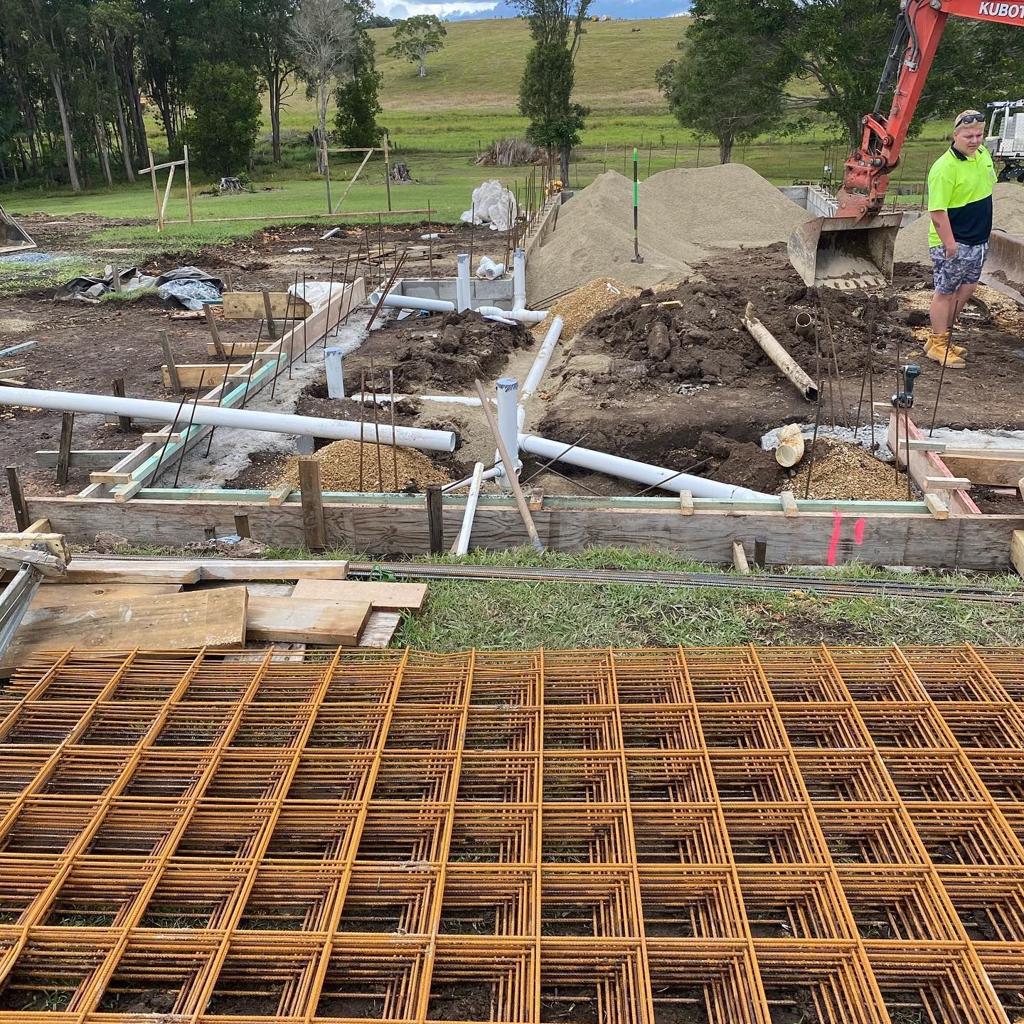 Construction site with plumbing, rebar, and a worker by an excavator. Green grass and dirt visible. — Daniel Gray Plumbing in Tyalgum, NSW