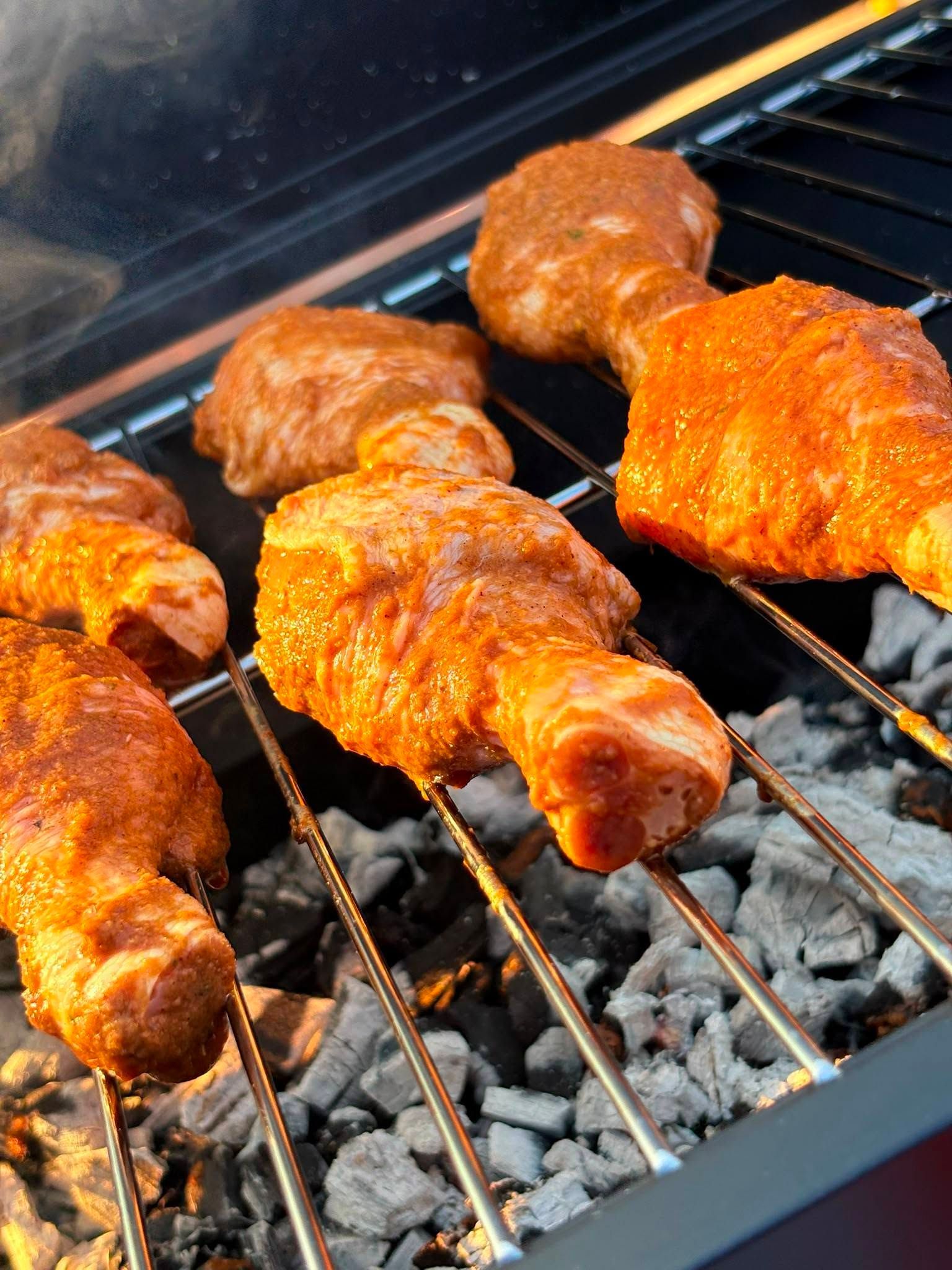 Chicken drumsticks grilling over hot coals on a barbecue, coated in a bright orange sauce.
