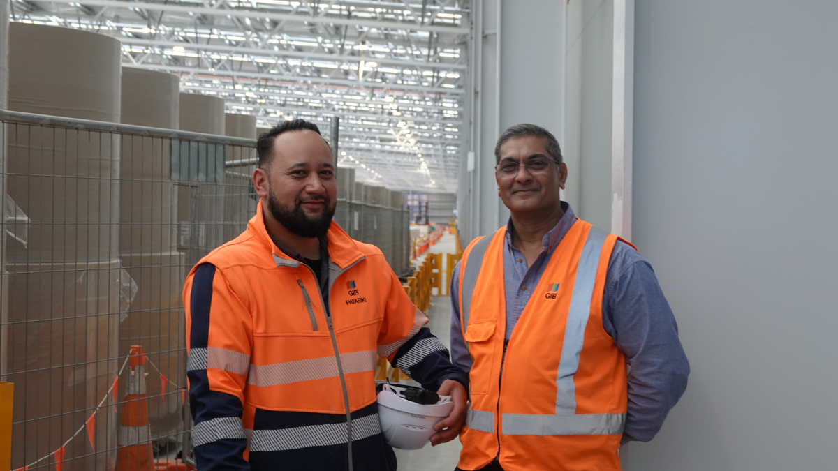 Two men in orange safety vests pose at a construction site. One holds a white hard hat, smiling.