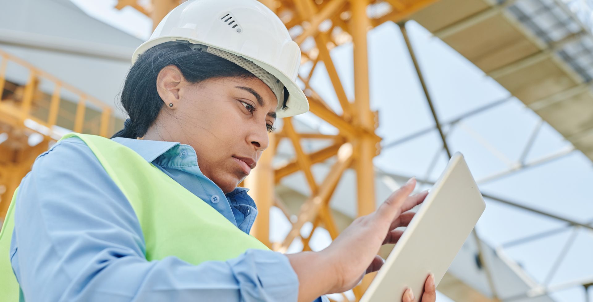 Woman in a hard hat and safety vest looking at a tablet at a construction site, crane visible in the background.