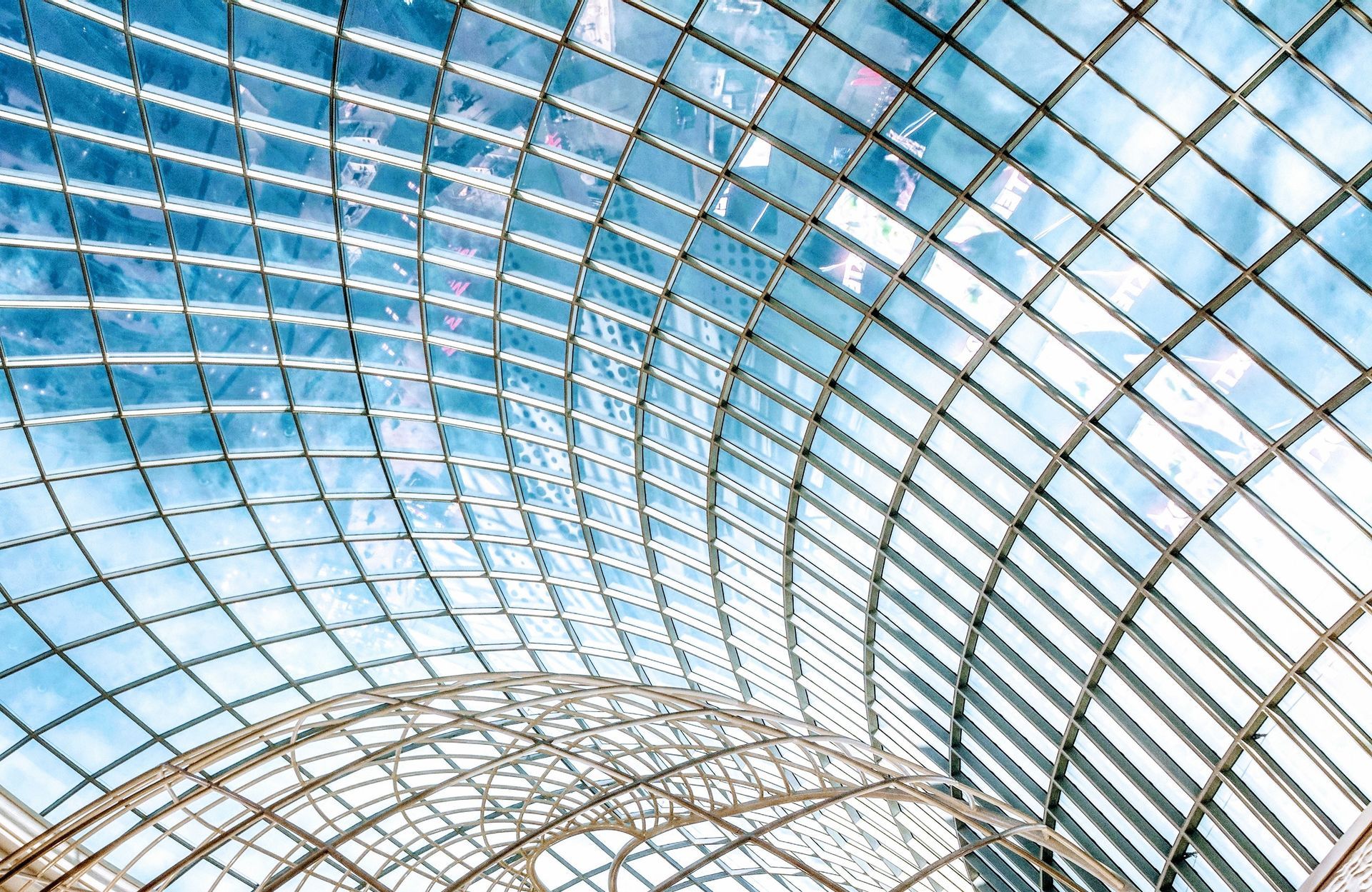 Curved glass roof with a grid pattern reflecting a bright blue sky. Steel framework supports the transparent structure.