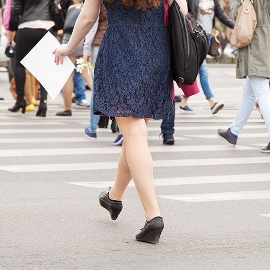 Woman in blue dress and wedge shoes walking across a crosswalk in a crowd, holding a blank white sign.