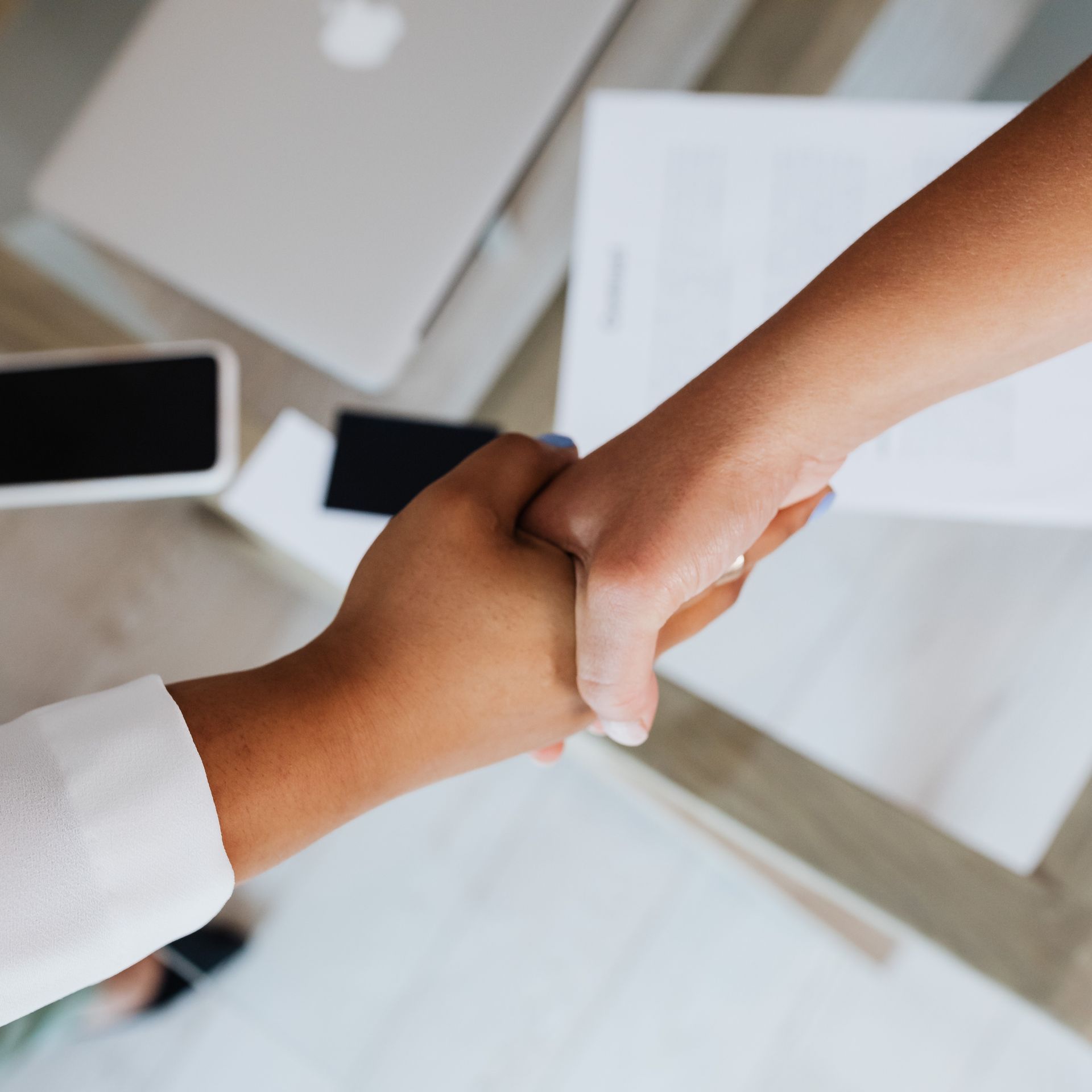 Two people shaking hands over a desk with a laptop and documents.