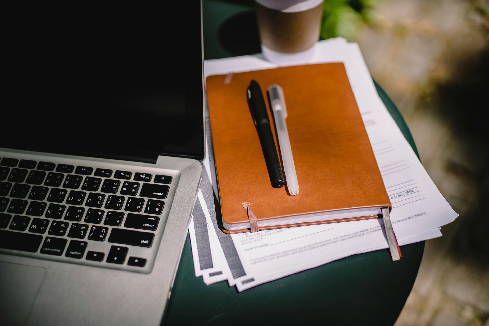 Brown Planner diary with two pens on top, next to an open laptop on a table
