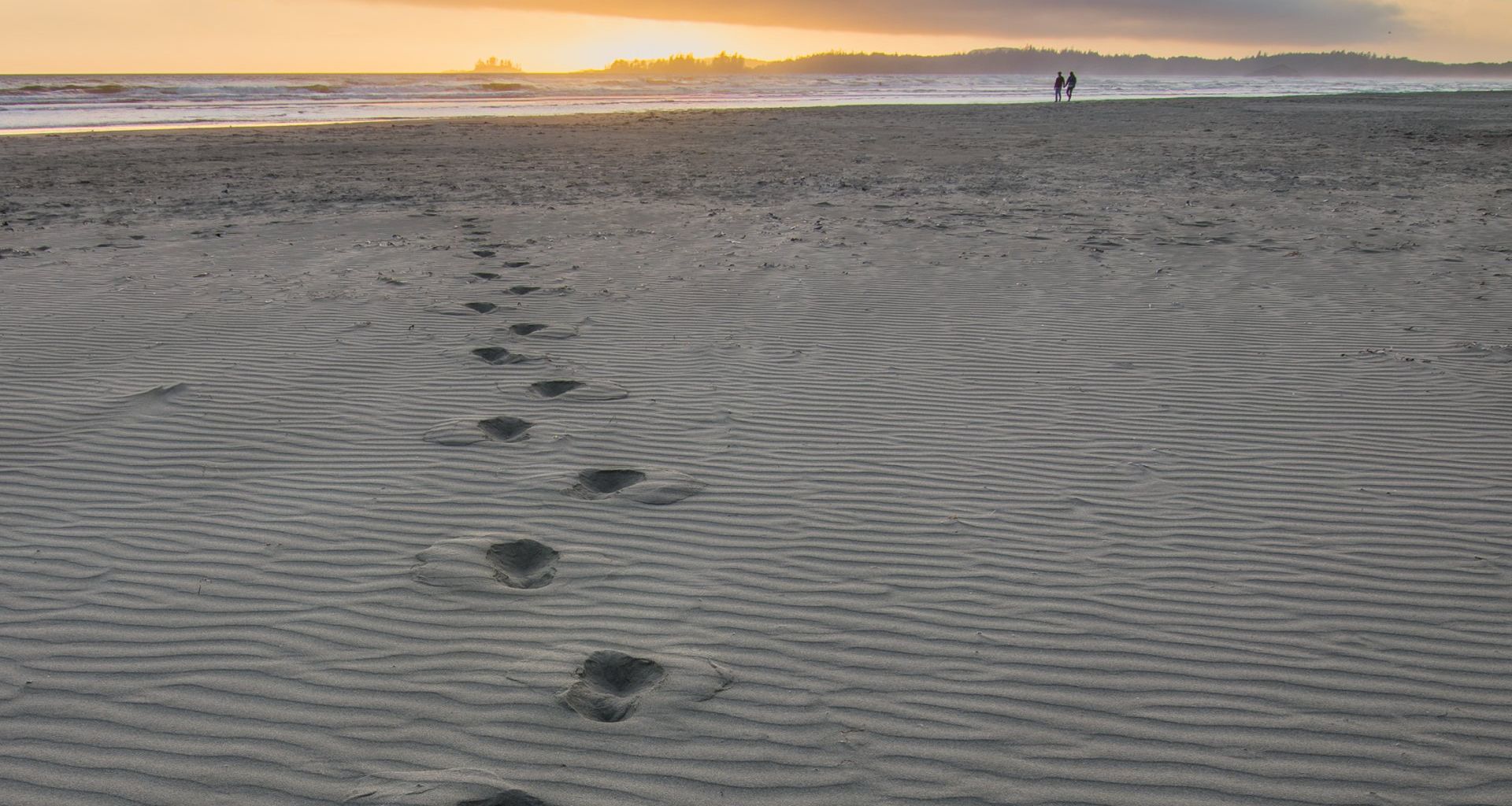 Footprints in sand lead toward two figures on a beach at sunset.