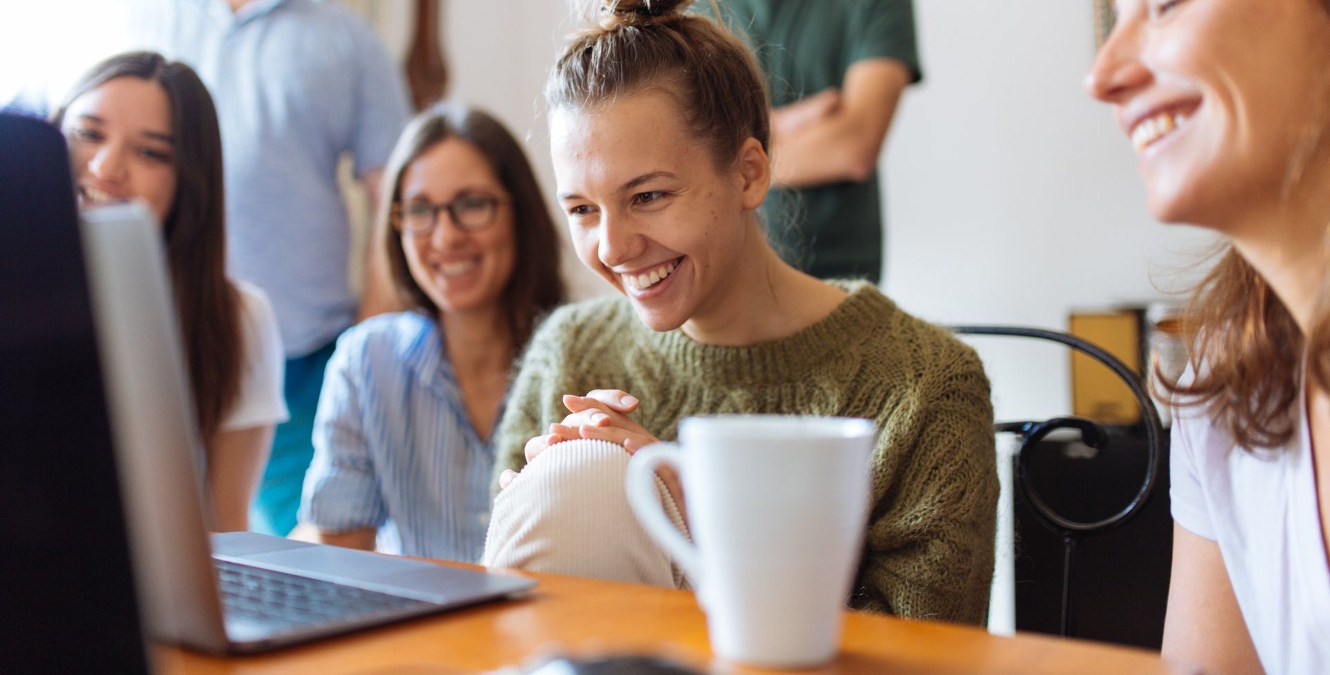 Group of people smiling and looking at a laptop on a table, possibly a collaborative workspace.