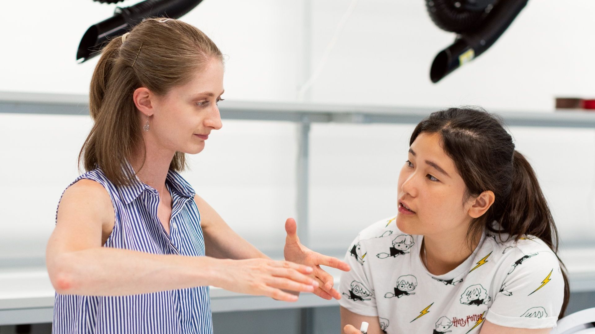 Two women in a lab setting, one gesturing with hands, the other listening intently. They are seated, discussing something.