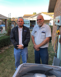 Two men standing outdoors beside a house and utility equipment, looking at the camera.